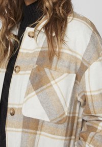 Kostkovaná vlněná bunda v krémové, hnědé a šedé barvě. Obsahuje velké knoflíky a náprsní kapsu s jemně kartáčovanou texturou.