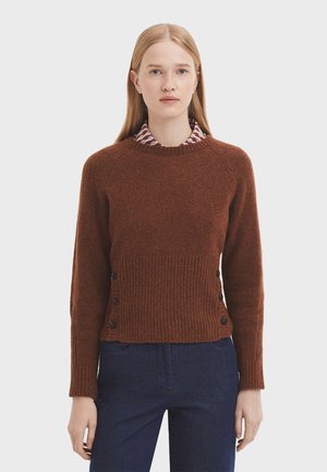 Brown wool sweater with ribbed texture, round neckline, and black buttons on the sides, worn over a collared shirt with a geometric pattern.