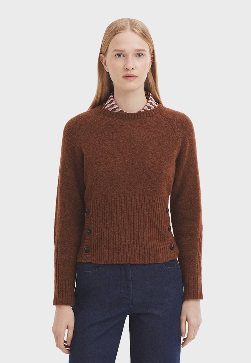 Brauner Wollpullover mit geripptem Textur, runder Ausschnitt und schwarzen Knöpfen an den Seiten, getragen über einem Kragenhemd mit geometrischem Muster.