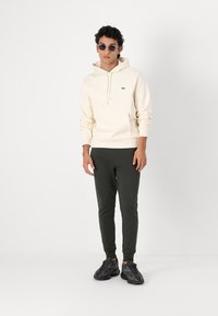 Haut à capuche crème avec un cordon de serrage, petit logo vert, associé à un pantalon slim-fit vert foncé et des baskets noires texturées. Le modèle porte des lunettes de soleil.