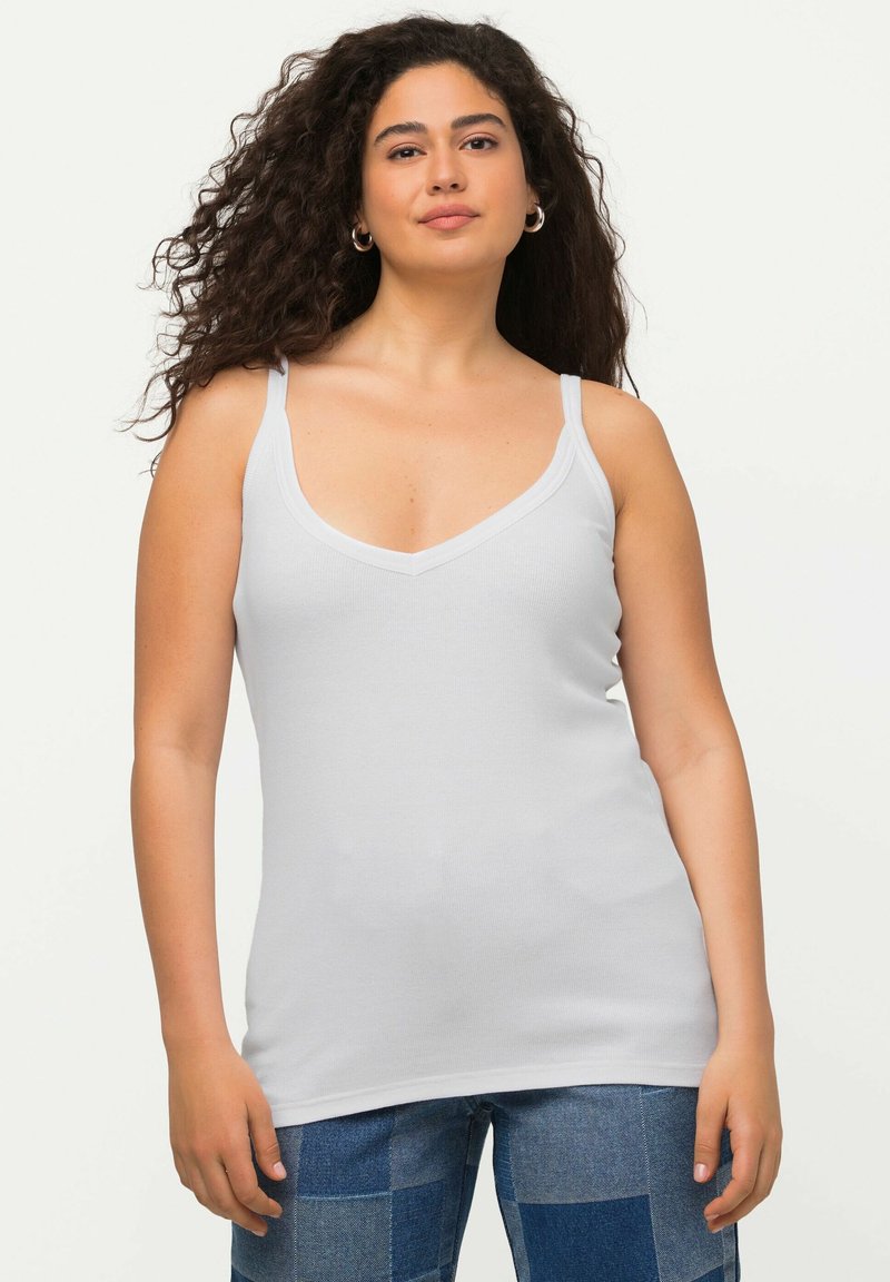 Top bianco senza maniche con spalline regolabili e scollatura a V; realizzato in tessuto liscio ed elasticizzato; abbinato a jeans blu con toppe.