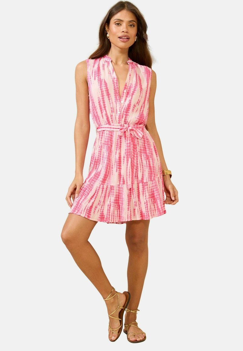 Abito rosa a maniche corte con vita legata, caratterizzato da un tessuto con un motivo tie-dye e gonna svasata. Indossato con sandali dorati e gioielli.