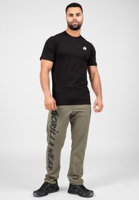 Zwarte T-shirt met korte mouwen en een wit logo, gecombineerd met olijfgroene trainingsbroek met zwarte grafische tekst langs één been. Zwarte sportschoenen.