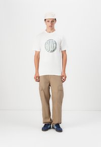 Hvit bomulls-t-skjorte med en rund grafisk trykk, kombinert med beige cargobukser med lomme på lapp, og blå sko med striper.