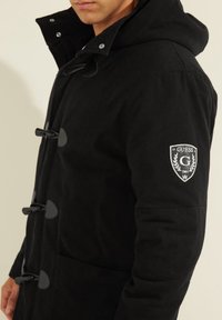 Parka negra con capucha, con cierre de pasador, bolsillos laterales y un logo de parche en la manga. Confeccionada con una tela texturizada.