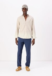 Camisa de lino de color beige claro, pantalones azul marino y zapatillas color tostado; la camisa tiene botones, mangas enrolladas y un corte relajado. La gorra añade un toque informal.
