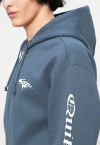 Tamno plavi dukserica na zip s kapuljačom od mekog pamuka; sadrži bijeli grafički motiv plamena na prsima i tekstualni dizajn koji se proteže niz rukav.