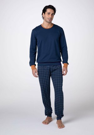 Uomo in piedi a piedi nudi che indossa una maglia a maniche lunghe blu navy con polsini e colletto arancioni, e pantaloni a quadri blu navy con linee arancioni a griglia.