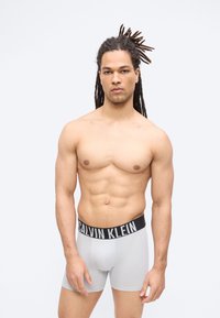 Μυώδης άνδρας με κοτσίδες στέκεται πρόσωπο, φορώντας ανοιχτό γκρι μποξέρ Calvin Klein, πάνω σε απλό λευκό φόντο.