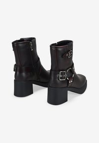 Botas de cuero marrón hasta el tobillo con un tacón bloque grueso, hebillas plateadas y un cierre lateral con cremallera. Textura suave con detalles en costura roja.