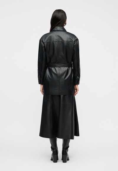 Γυναίκα με μακριά σκούρα μαλλιά, φοράει μαύρο δερμάτινο μπουφάν με ζώνη, ταιριαστό δερμάτινο μίντι φούστα και μαύρες μπότες μέχρι το γόνατο, φανερή από πίσω.