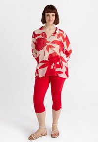 Blouse florale rouge avec un fond clair, dotée d'un col en V et de manches longues. Assortie avec un pantalon rouge court et des sandales beiges.