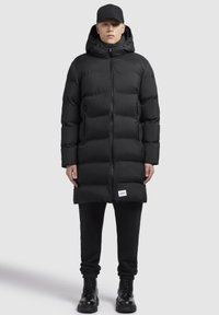 Sort pufferjakke med hætte, med quiltet design, lynlås lukning og forlommer. Båret over sorte sweatpants og snørebåndsstøvler.