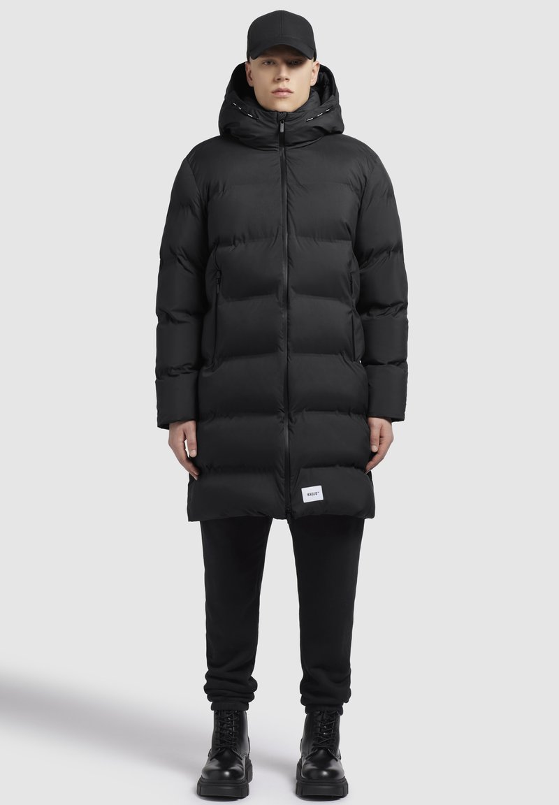 Sort pufferjakke med hætte, med quiltet design, lynlås lukning og forlommer. Båret over sorte sweatpants og snørebåndsstøvler.