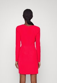 Kvinna med flätad hästsvans iförd en passande långärmad klänning i klarr rosa, stående vänd mot en vit bakgrund.