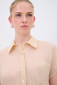 Camicia button-up color pesca chiaro con tessuto texture, maniche corte, colletto e tasca a toppa. La camicia presenta sottili righe orizzontali.