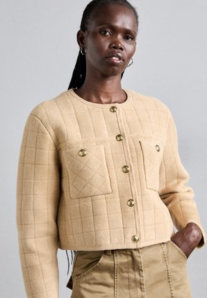 Femeie cu părul împletit, purtând o jachetă scurtă bej, matlasată, cu nasturi aurii și pantaloni bej cu talie înaltă, cu o mână în buzunar.