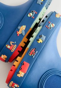 Blauwe rubberen schoen met een structuuroppervlak, versierd met kleurrijke voertuigapplicaties: vliegtuigen, vrachtwagens en een helikopter in oranje, rood, geel en groen.