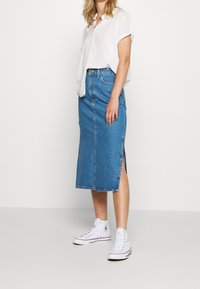 Denim-mittapituinen hame sivuhalkiolla ja keskisaumalla, yhdistetty valkoiseen lyhythihaiseen paitaan ja valkoisiin korkeavartisiin tennareihin.