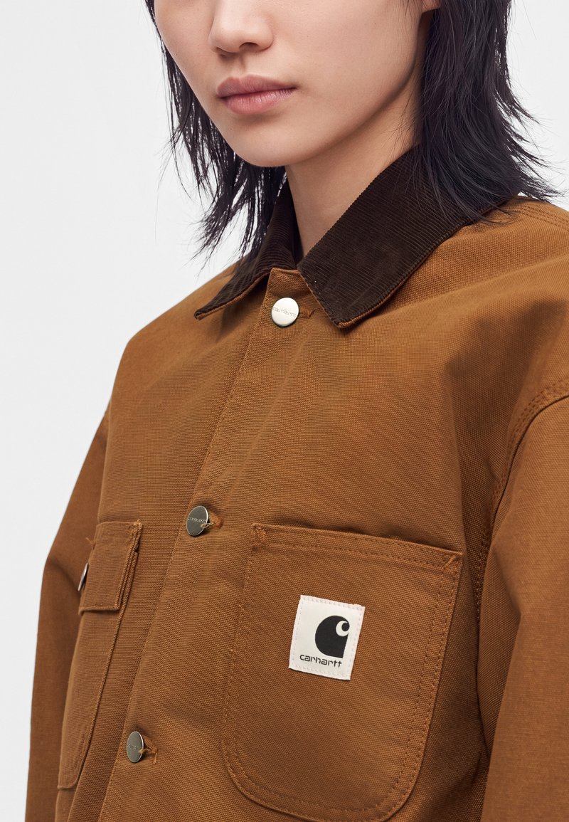 Person wearing a brown Carhartt jacket with dark collar and visible front pockets and metal buttons, shown from the shoulders to mid-chest.
