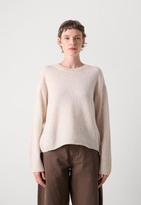 Lys beige strikkegenser med rund hals, ribbestrikkede mansjetter og en avslappet passform, kombinert med løse brune bukser. Myk tekstur synlig.