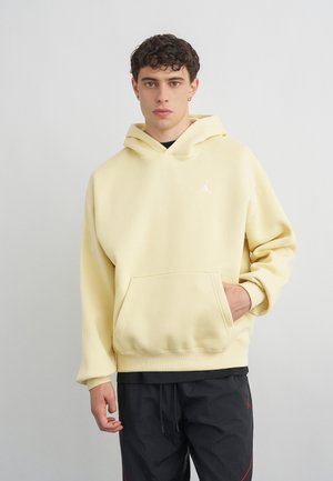 Mlad moški s kratkimi kodrastimi lasmi, ki nosi svetlo rumeno kapucarico z žepom spredaj in črne hlače na enostavni sivi podlagi.