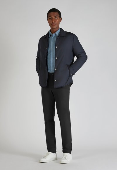 Marineblauwe pufferjacket met drukknoopsluitingen, gedragen over een lichtblauw overhemd met knopen, gecombineerd met zwarte broek en witte sneakers.