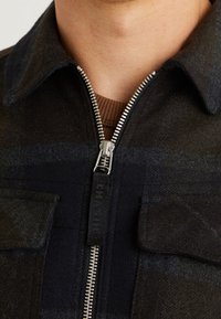 Detailní záběr tmavé kárované bundy se stříbrným zipem, částečně zapnutým přes hnědý žebrovaný límec košile.