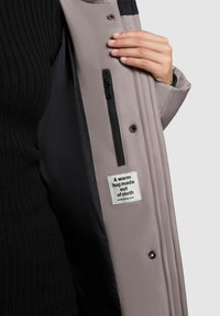 Lys grå pufferjakke med en ribbet sort indvendig, der har en lynlåslomme og et label, der står "Et varmt kram lavet af stof."