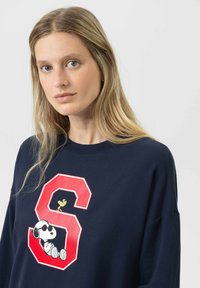 Blonde vrouw met blauwe ogen in een marineblauwe sweatshirt met een grote rode "S", met Snoopy in een zonnebril en een Woodstock-vogel erbovenop.