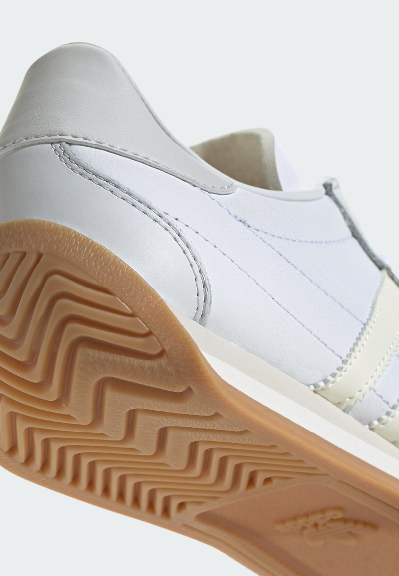Zapatilla de cuero blanca con detalles en gris y costuras. Suela de goma con un patrón de tracción en zigzag. Diseño de tacón redondeado.
