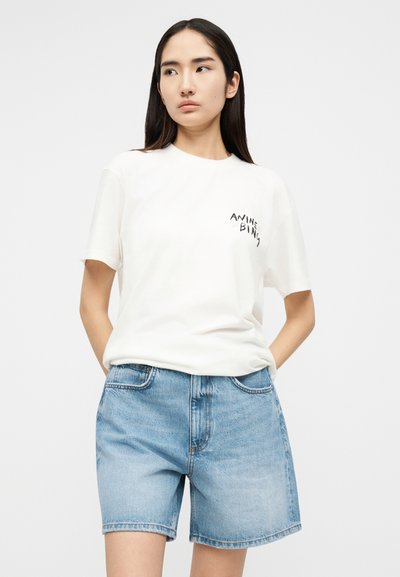 Hosszú sötét hajú nő fehér rövid ujjú pólót visel felirattal és világoskék farmer rövidnadrágot, zsebeiben tartja a kezét.