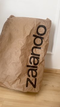 Smeđi zgužvani papirnati vrećica s velikim crnim logotipom "Zalando", koja ima jednostavan, pravokutni oblik i teksturiranu površinu na svijetlom drvenom podu.