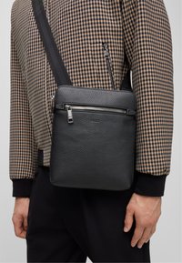 Czarna skórzana torba na ramię z fakturowaną powierzchnią, srebrnym zamkiem błyskawicznym i regulowanym paskiem. Nosi się ją na tle kurtki w kratkę.