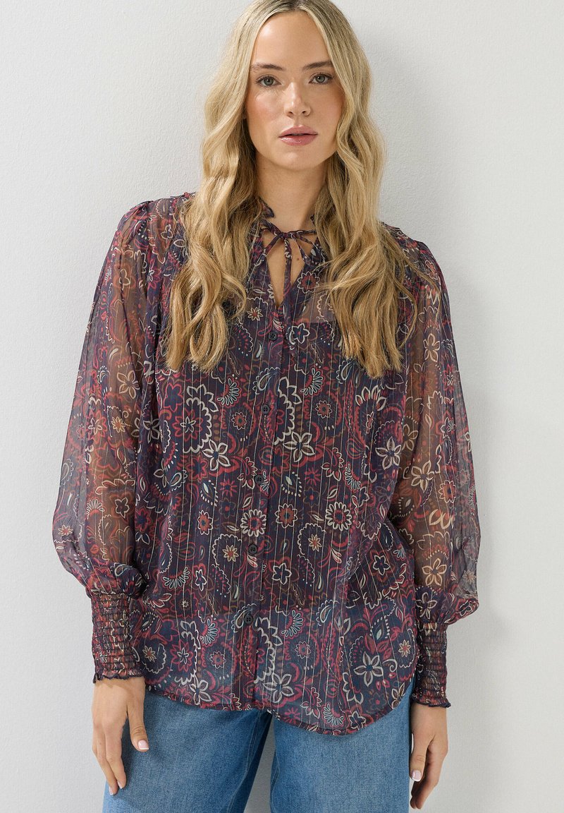 Transparante blouse met bloemenpatroon op een donkere achtergrond, met een strikbij de hals en gathered cuffs. Gepaard met lichtblauwe spijkerbroek.