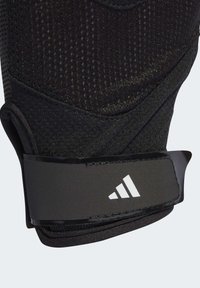 Svart atletisk stöd med ett texturerat mesh-tyg, vadderade områden och ett brett band med en vit logotyp. Släta metalldetaljer ingår.
