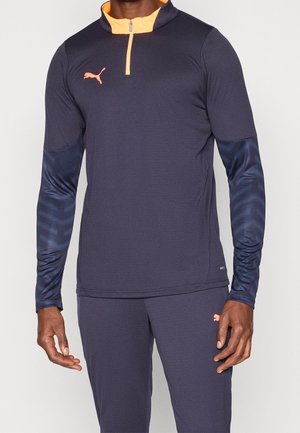 Mann trägt dunkelblaue Puma-Sportbekleidung mit langen Ärmeln sowie orangefarbenem Kragen und Logo, steht vor einem einfarbigen Hintergrund.