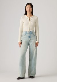 Κρεμ ριμπ πλεκτό τοπ με μακριά μανίκια και λεπτομέρειες με κουμπιά, συνδυασμένο με τζιν ελαφριάς πλύσης, φαρδιάς γραμμής. Ομαλή υφή, άνετη εφαρμογή και κλασικός σχεδιασμός.