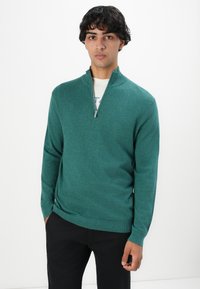 Junger Mann mit lockigem, dunklem Haar, der einen tealfarbenen Half-Zip-Pullover über einem weißen Hemd und schwarzen Hosen trägt, steht vor einem schlichten weißen Hintergrund.