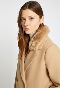 Camelkleurige jas met een nepbontkraag, dubbele knopen, gladde textuur en grote knopen. Gestyled met een lichte coltrui eronder.
