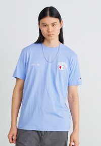 Gaiši zils kokvilnas T-krekls īsām piedurknēm, ar sarkanā un baltā Champion logo uz krūtīm un nelielu baltu teksta detaļu.
