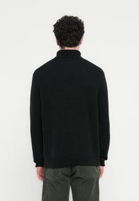 Μαύρο πουλόβερ με ψηλό λαιμό, άνετη γραμμή και ριγέ μανσέτες, φτιαγμένο από πλεκτό ύφασμα· φορεμένο με παντελόνι από κοτλέ σε σκούρο ελαιδί χρώμα.