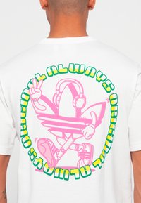 Pessoa vestindo uma camisola branca com um personagem de tênis de cartoon rosa usando auscultadores e fazendo um sinal de paz, rodeada de texto colorido.