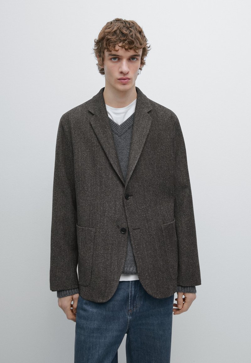 Giovane uomo con capelli ricci indossa un blazer di lana marrone sopra un maglione grigio e una camicia bianca, abbinati a jeans blu, in piedi davanti a uno sfondo semplice.