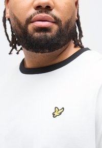 Άνδρας με γενειάδα που φορά λευκό πουκάμισο με μαύρο γιακά και μικρό κίτρινο πουλί ως έμβλημα στο στήθος.