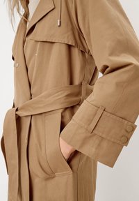 Beige trenchcoat med bälte i midjan, sidofickor och knäppningar vid ärmsluten, tillverkad av slätt, lättviktigt tyg med en strukturerad design.