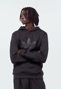 Young man with dreadlocks wearing a black Adidas hoodie with arms crossed, looking to the side against a plain light background.