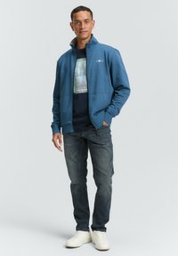 Blå zip-op jakke med høj krave, lagd over en marineblå grafisk t-shirt. Sammen med mørke jeans og lyse sneakers, med en afslappet pasform.