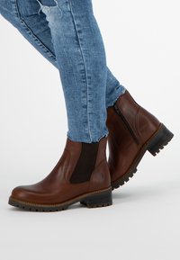 Hnědé kožené kotníkové boty s elastickými bočními panely, kulatou špičkou a robustní gumovou podrážkou. Nesené s světle modrými džíny.