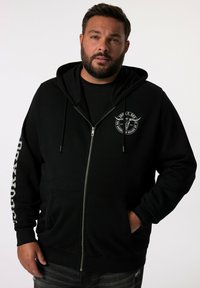 Sudadera con cremallera negra hecha de un tejido suave, con un parche de logo en el frontal y bolsillos laterales. Mangas largas con detalles de texto en uno de los brazos.